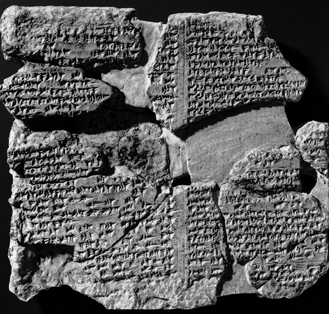 Image en noir et blanc représentant une tablette en argile fragmentée, couverte d'inscriptions cunéiformes. Les morceaux de la tablette semblent usés et cassés, laissant apparaître des textes anciens gravés, probablement issus d'une civilisation mésopotamienne.